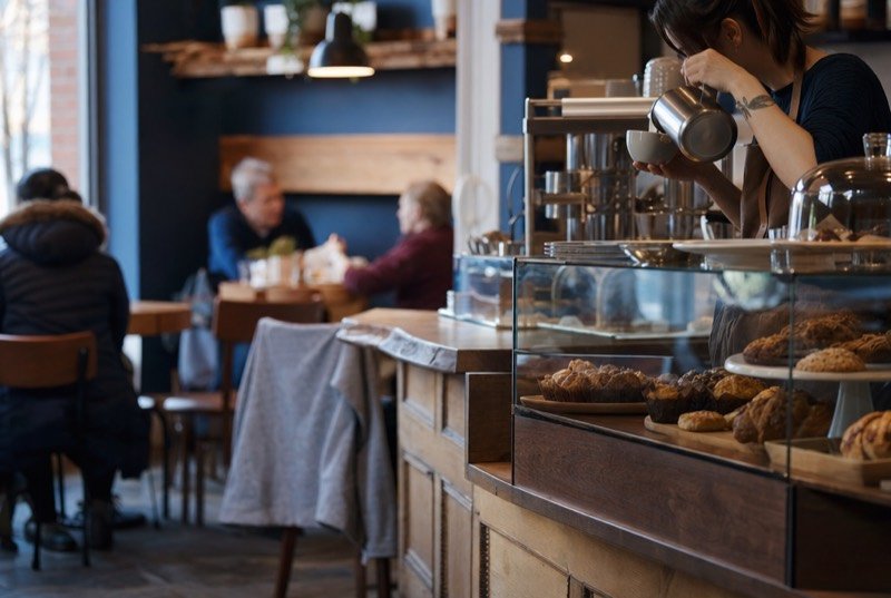 A café counter with fresh baking under glass and a barista pouring coffee