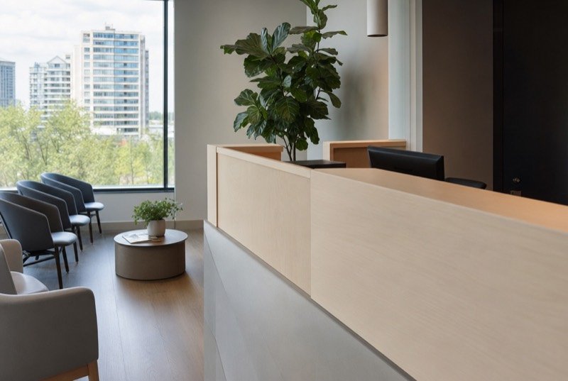 A clinic front desk in pale wood beside a tall, bright window
