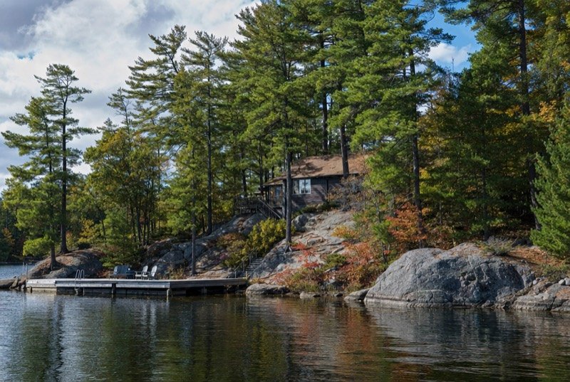 A wooden cottage and dock on a granite shoreline among white pines, with calm water in early autumn