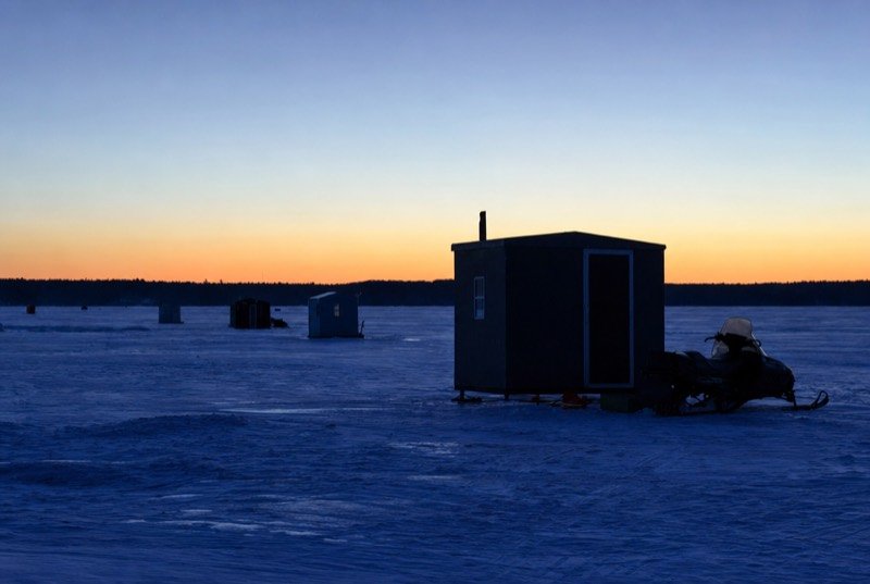 Ice fishing huts on a frozen lake at sunrise, a snowmobile parked beside one