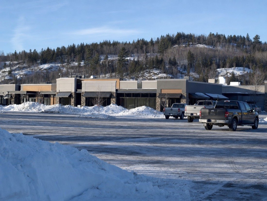 A one-storey plaza of small shops with plowed parking, snowbanks and pine-covered hills behind
