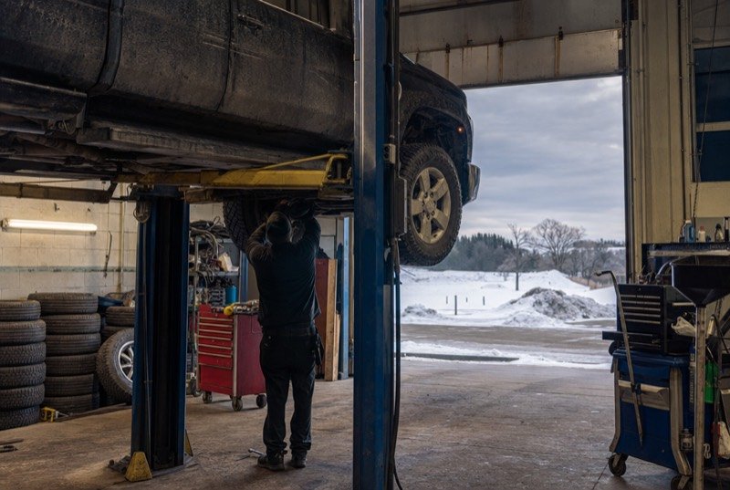 A pickup raised on a garage hoist with winter tires stacked along the wall